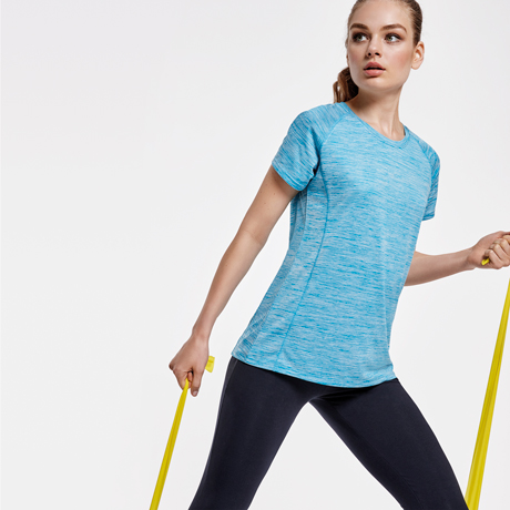 Mujer con camiseta deportiva azul claro de manga corta y pantalones negros haciendo ejercicio con banda elástica amarilla.