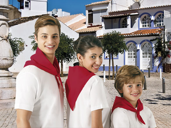 Tres personas con pañuelos rojos y camisetas blancas en una plaza con casas blancas y tejados rojos.